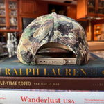 Camouflage-patterned hat on top of a book titled 'Wanderlust USA' with a blurred indoor background.