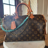 Designer handbag with monogram pattern on a surface adorned with Turquoise beaded bag chain with gold chain on a wooden block, J Forks & Leather and Vodka
