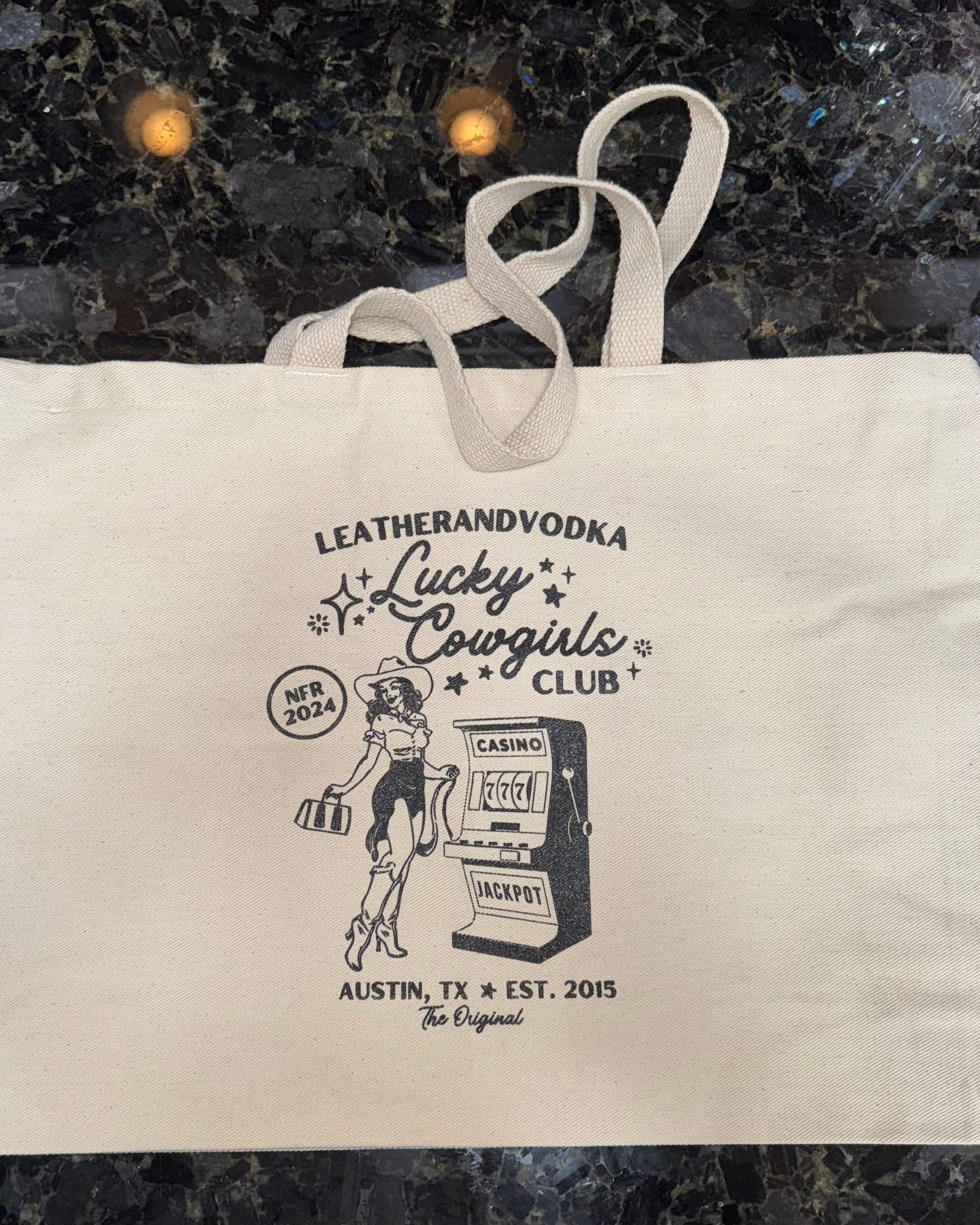 A beige cotton tote bag with a printed logo featuring an illustration of a cowgirl and a slot machine, text mentioning 'Leatherandvodka', 'Lucky Cowgirls Club', and 'Austin, TX'. The bag also includes the text 'NFR 2024' and has a graphic indicating a five-day event.