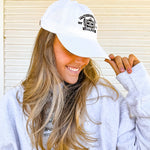 Woman wearing a white cap and light gray sweatshirt with a logo, standing in front of a white wall.