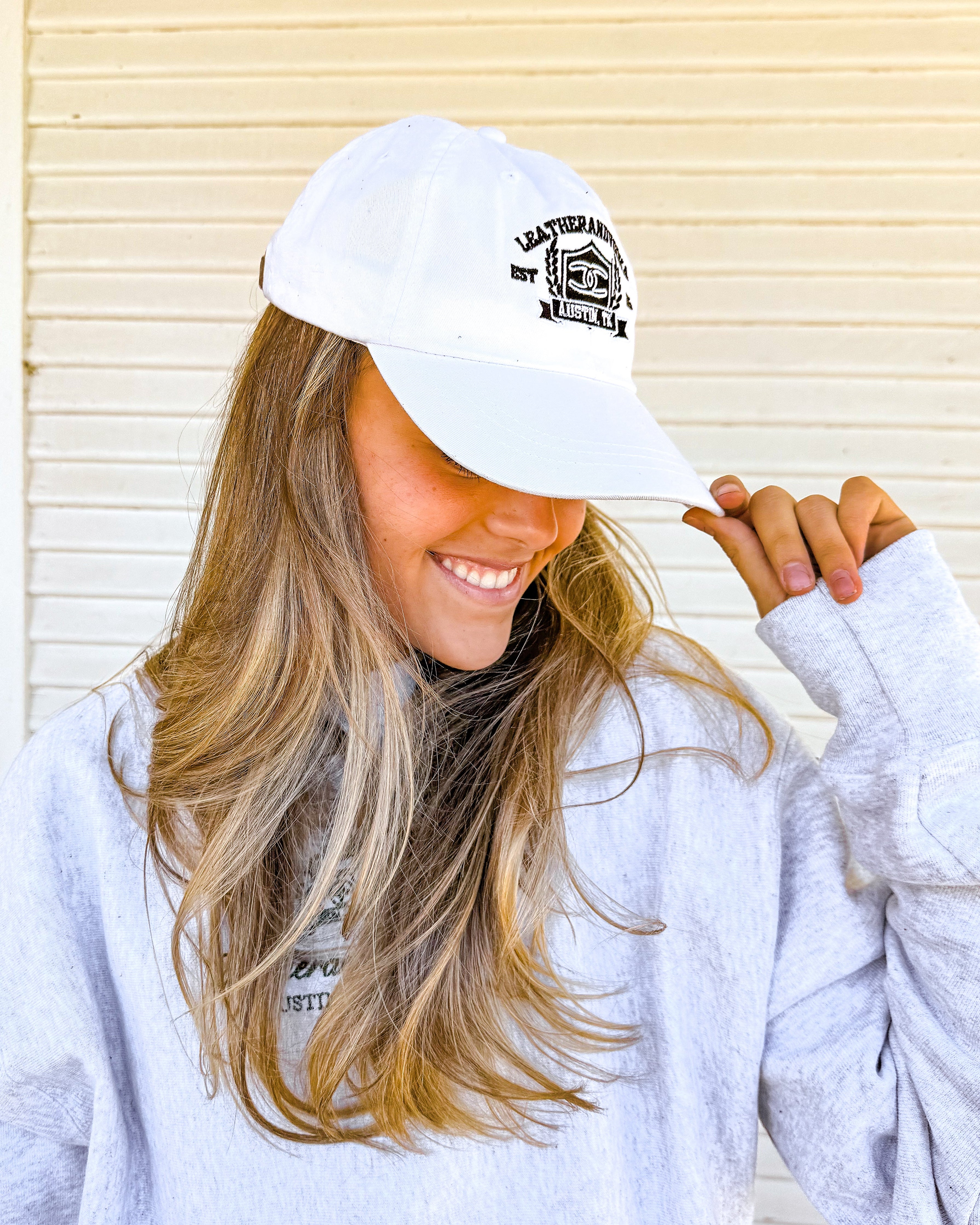 Woman wearing a white cap and light gray sweatshirt with a logo, standing in front of a white wall.