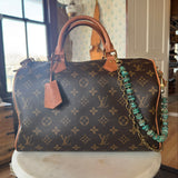 Designer handbag with monogram pattern on a surface adorned with Turquoise beaded bag chain with gold chain on a wooden block, J Forks & Leather and Vodka