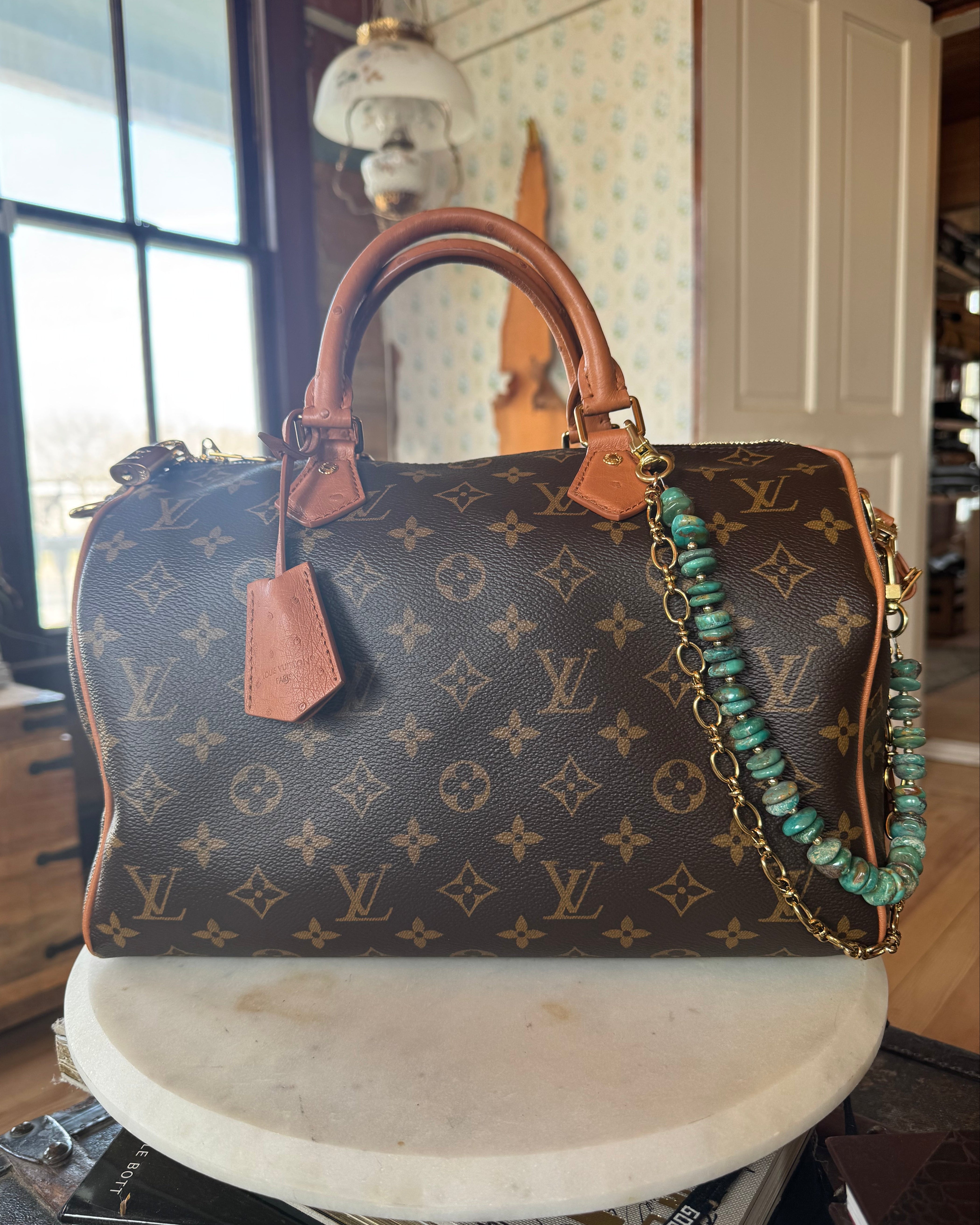 Designer handbag with monogram pattern on a surface adorned with Turquoise beaded bag chain with gold chain on a wooden block, J Forks & Leather and Vodka