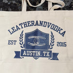 A beige cotton tote bag with 'LEATHERANDVOODKA' and 'AUSTIN, TX' printed in blue, along with a graphic with horseshoes.