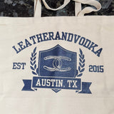 A beige cotton tote bag with 'LEATHERANDVOODKA' and 'AUSTIN, TX' printed in blue, along with a graphic with horseshoes.