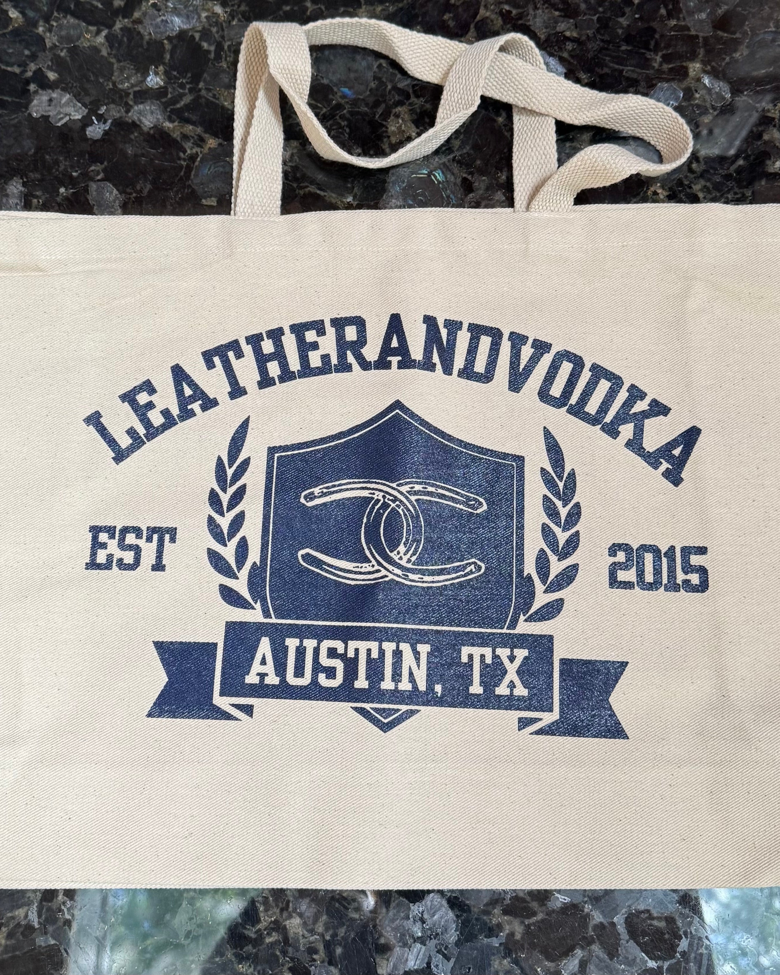 A beige cotton tote bag with 'LEATHERANDVOODKA' and 'AUSTIN, TX' printed in blue, along with a graphic with horseshoes.