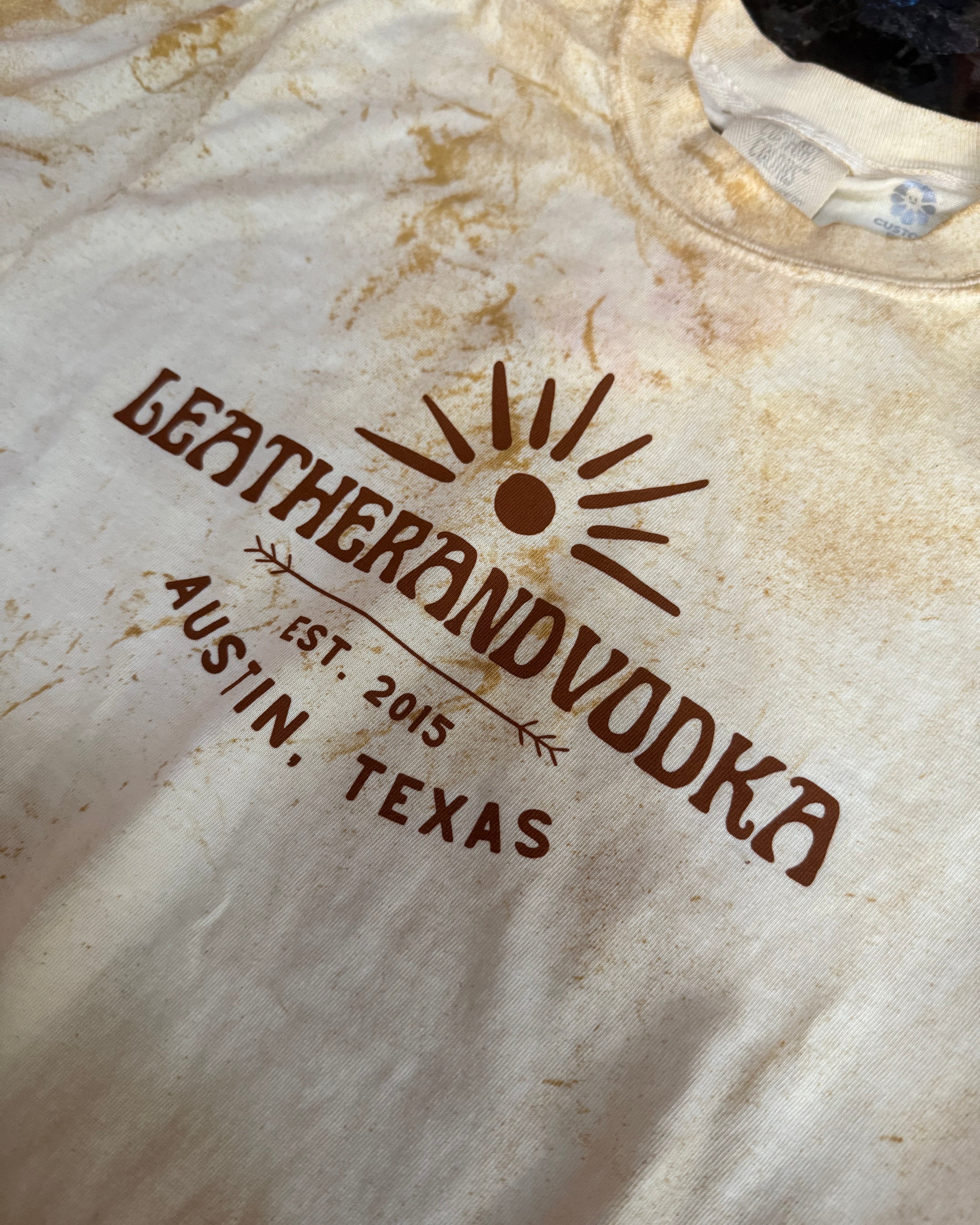 Leather and Vodka logo on a tye-dye t-shirt, laying flat on table.