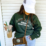 Person wearing a green LAV sweatshirt with text, holding a drink, and carrying a handbag against a white wall.