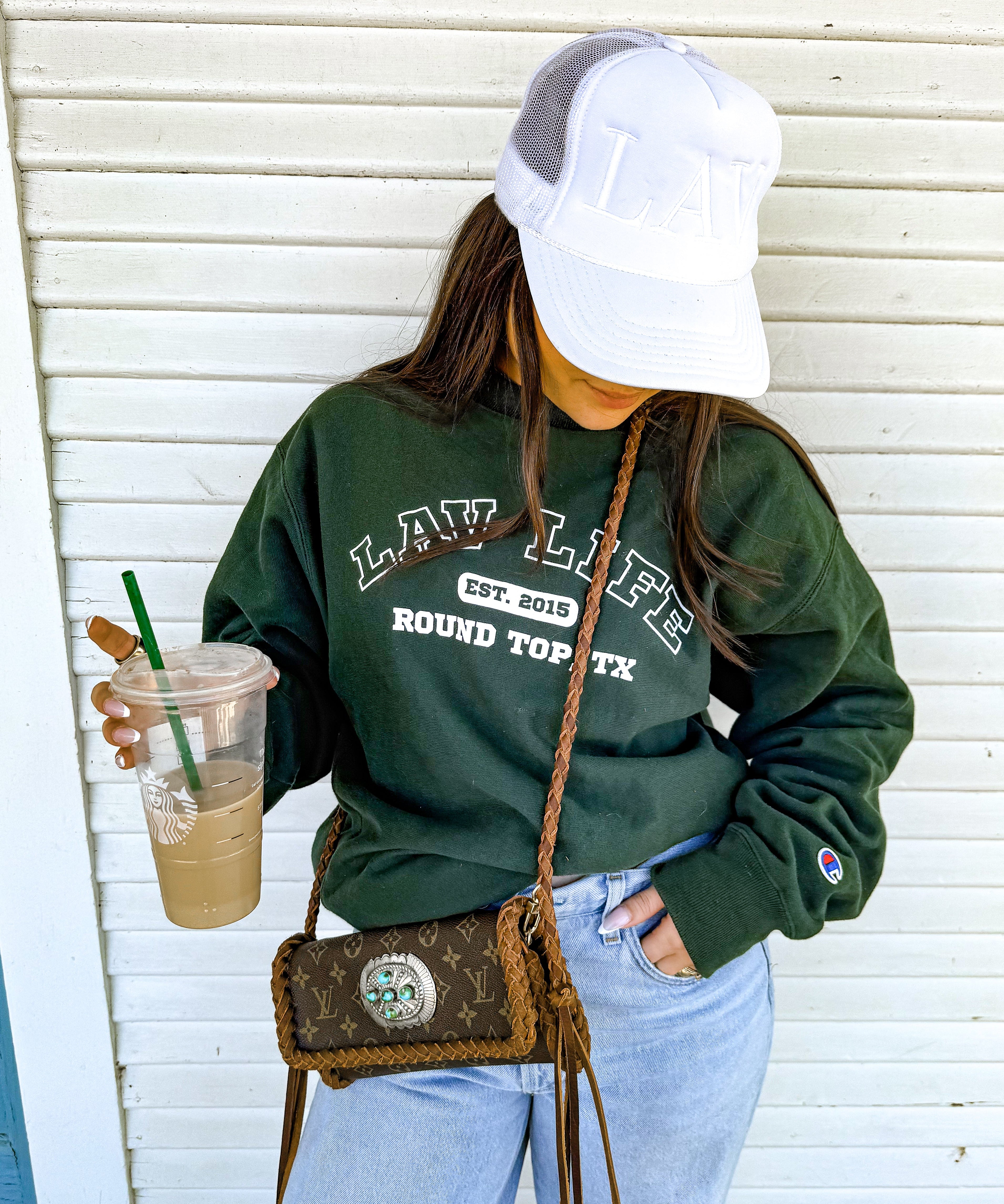 Person wearing a green LAV sweatshirt with text, holding a drink, and carrying a handbag against a white wall.