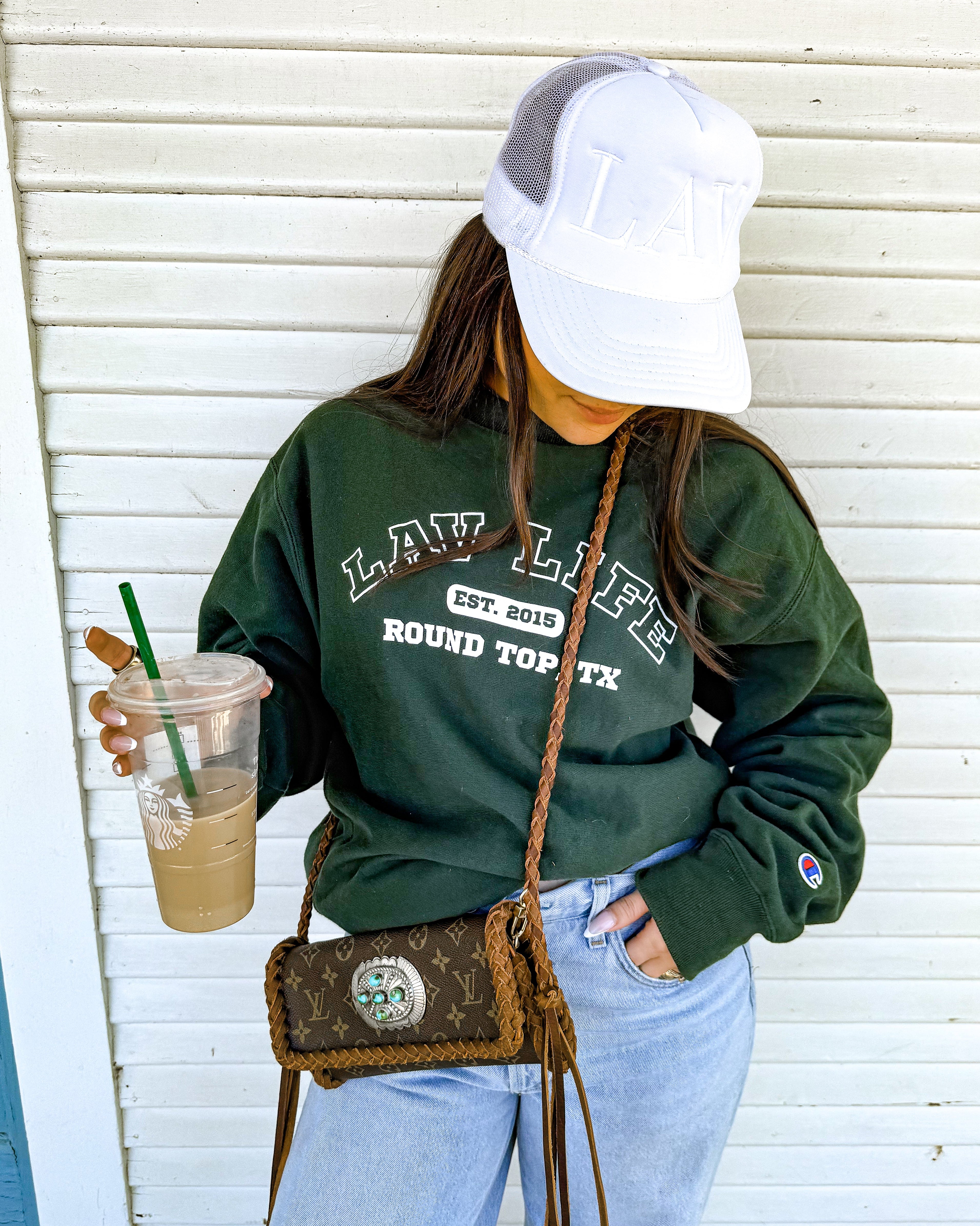 Person wearing a green LAV sweatshirt with text, holding a drink, and carrying a handbag against a white wall.