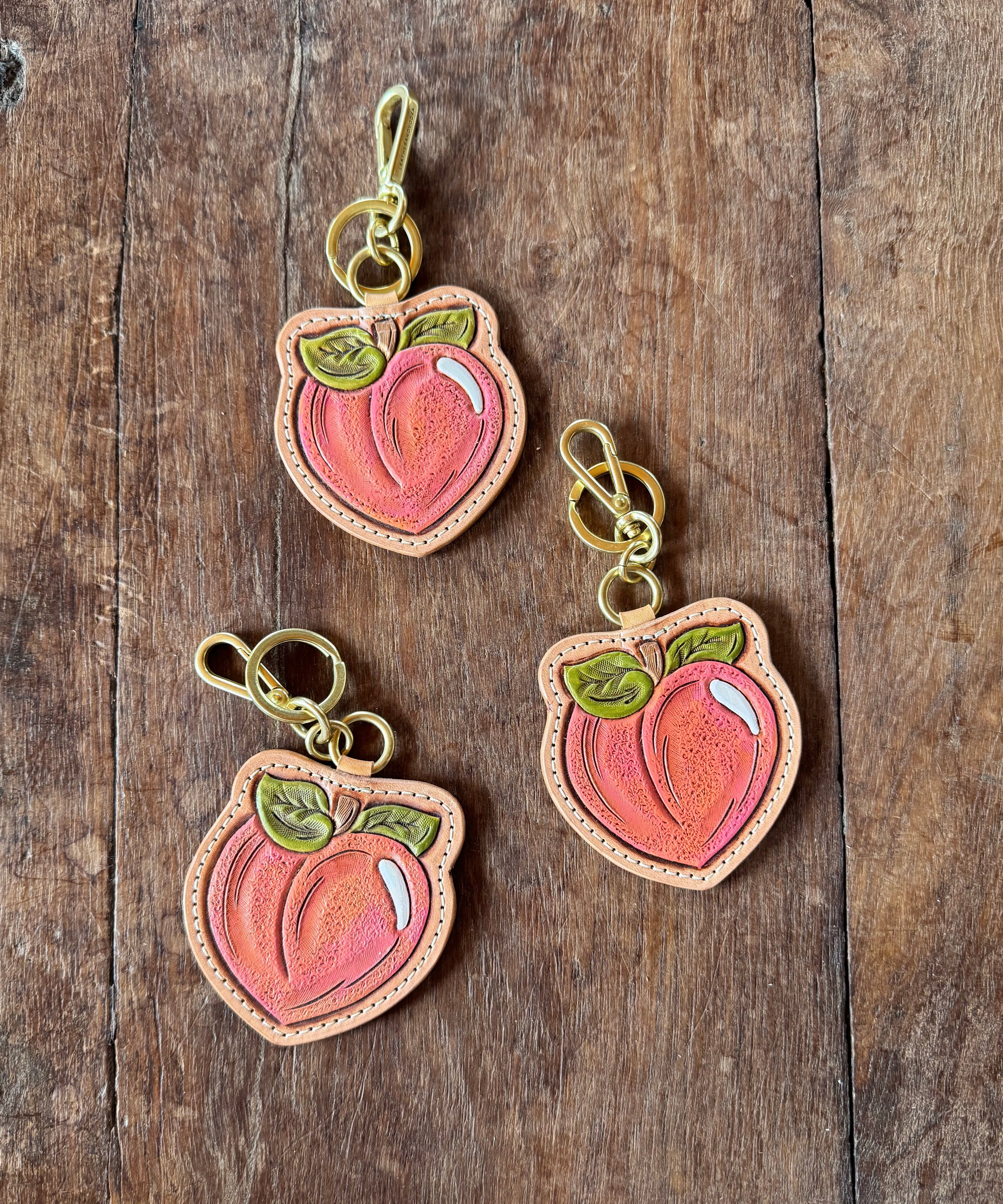 Three peach-shaped leather keychains, hand bag charms on a wooden surface. Collab between Leatheradvodka and ALLRed out of Round Top Texas
