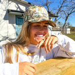 Person wearing a Leather & Vodka, Round Top Texa camouflage cap and white shirt outdoors on a sunny day.