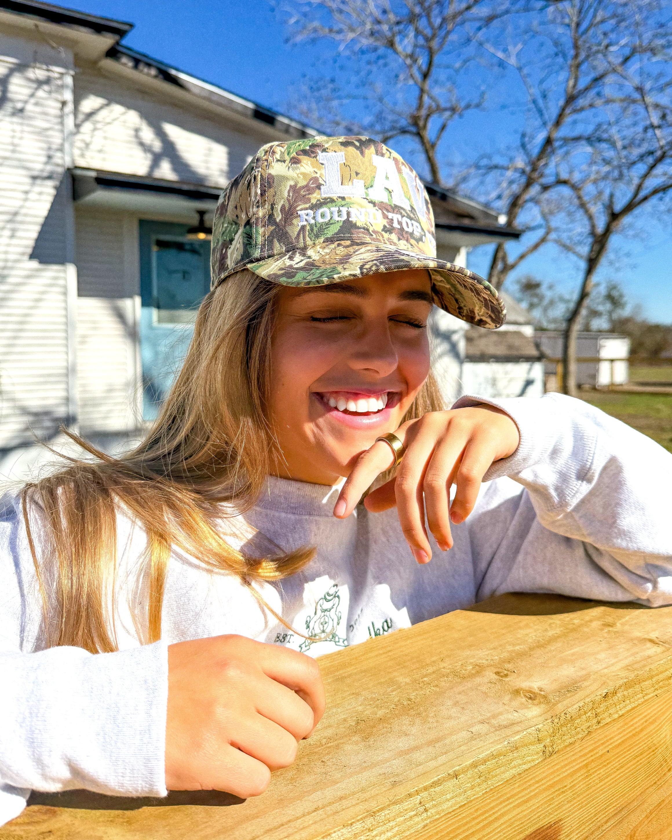 Person wearing a Leather & Vodka, Round Top Texa camouflage cap and white shirt outdoors on a sunny day.