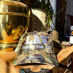 Camouflage cap with 'LAV Round Top, TX' text on a wooden surface with decorative items.