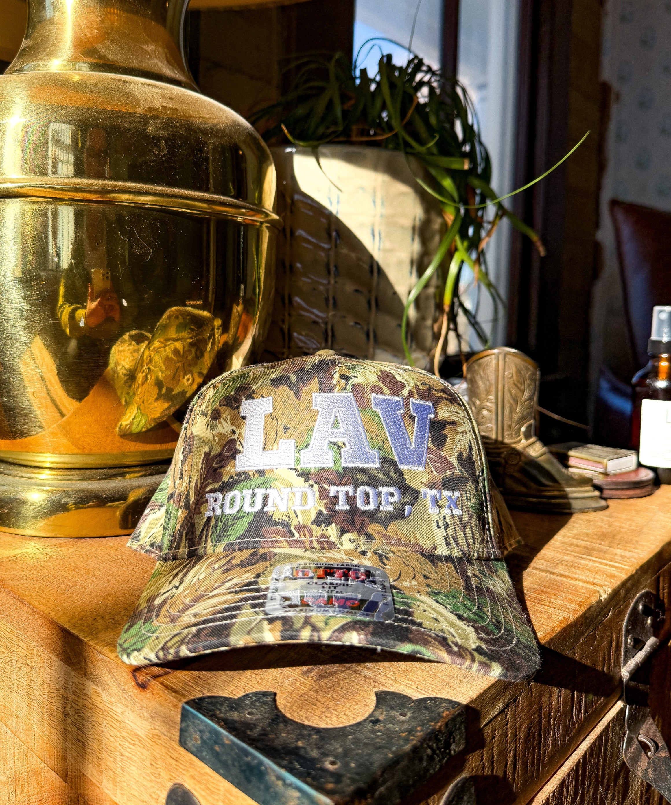Camouflage cap with 'LAV Round Top, TX' text on a wooden surface with decorative items.