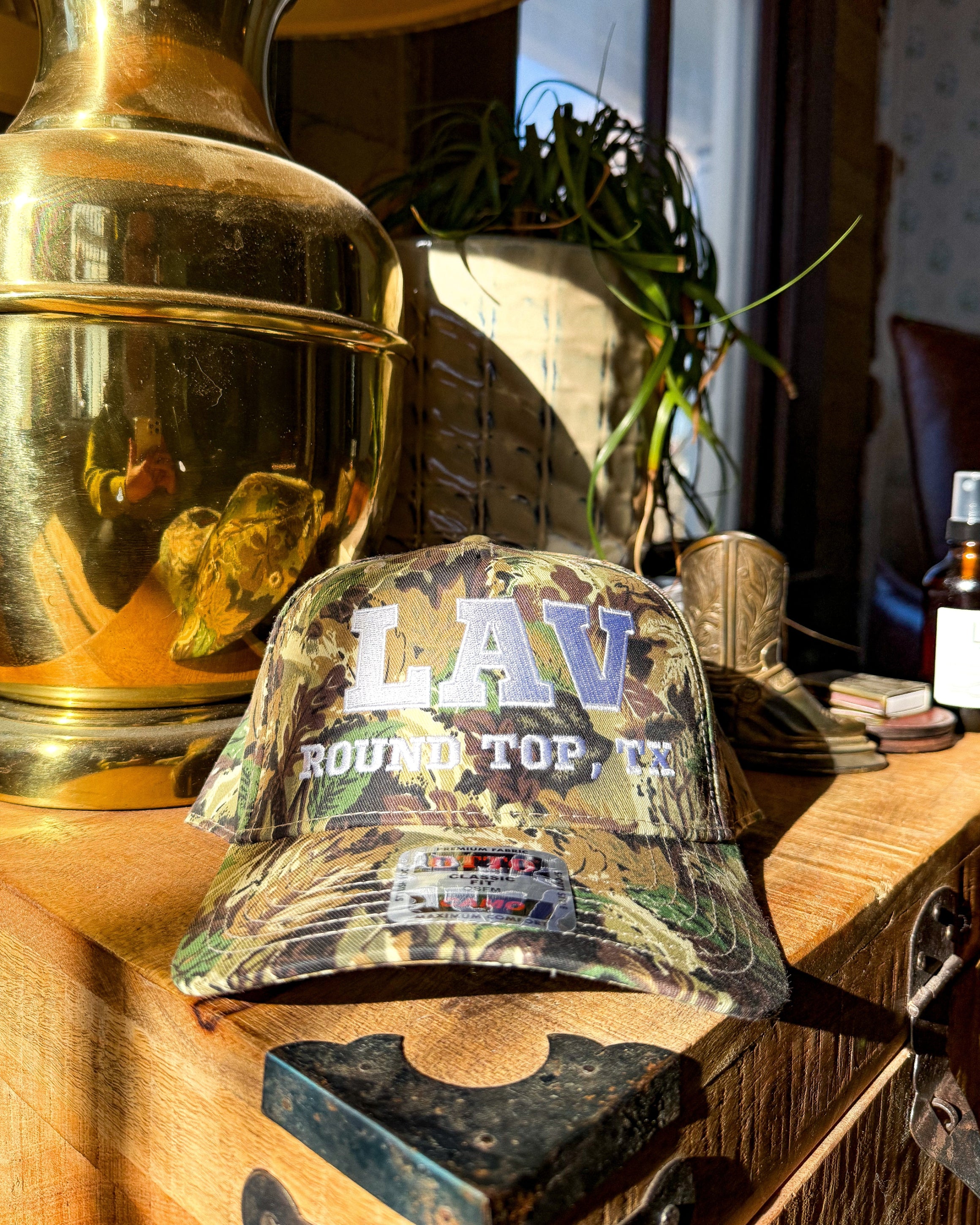 Camouflage cap with 'LAV Round Top, TX' text on a wooden surface with decorative items.