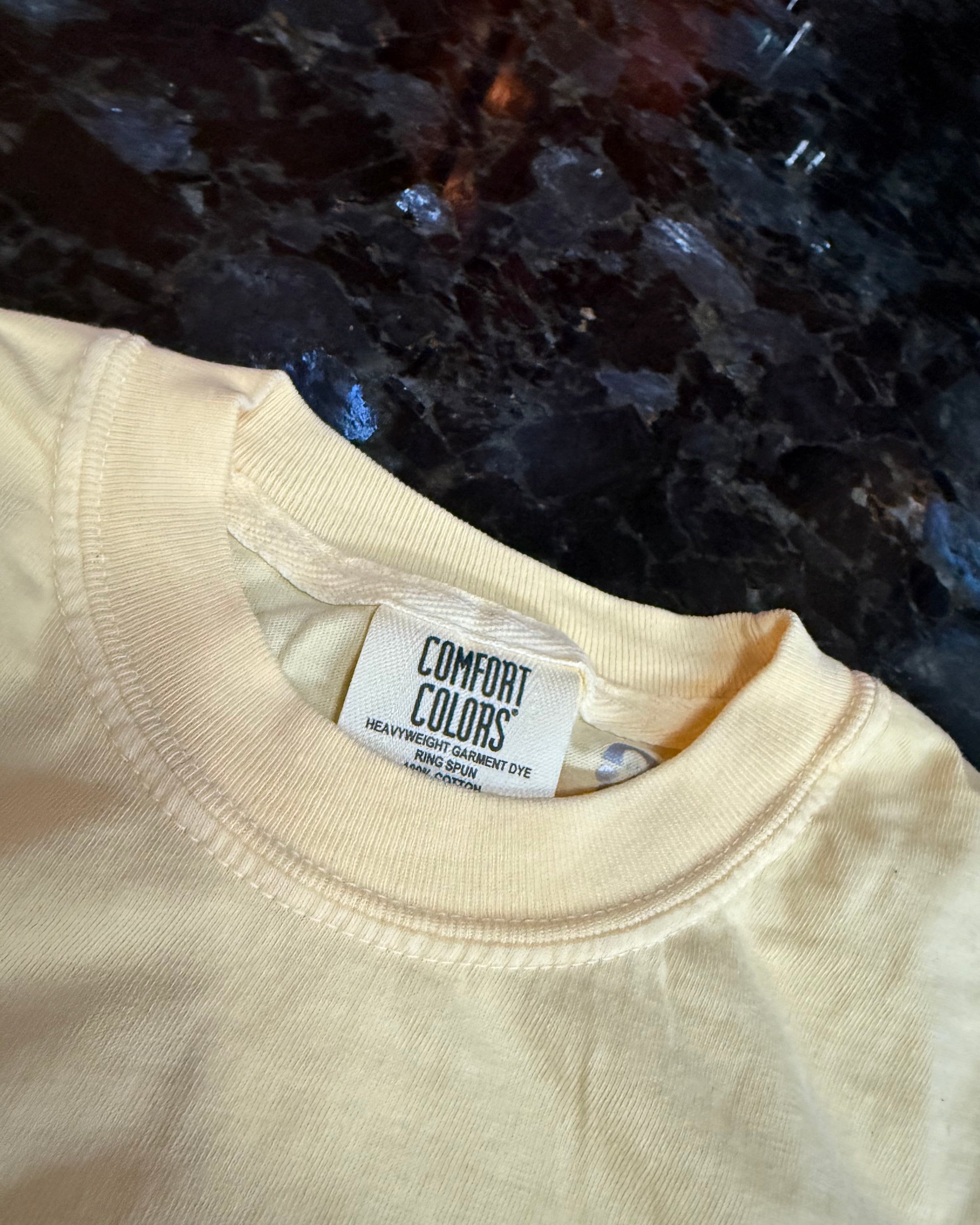 Beige t-shirt with a 'Comfort Colors' tag against a dark textured background