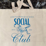 A beige cotton tote bag with 'LEATHERANDVOODKA SOCIAL CLUB 2015 ATX' printed in blue text on the front.