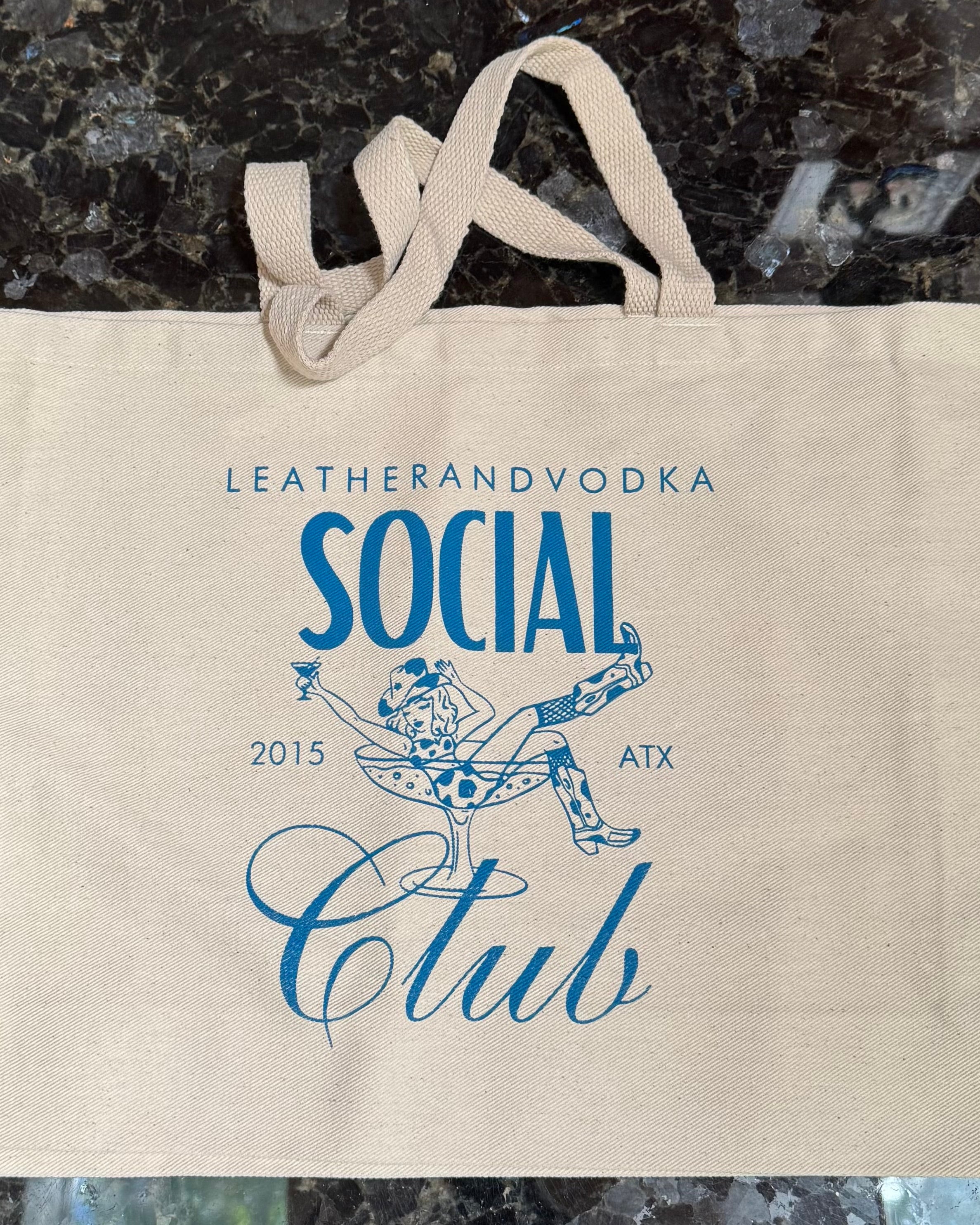 A beige cotton tote bag with 'LEATHERANDVOODKA SOCIAL CLUB 2015 ATX' printed in blue text on the front.