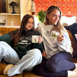 Two women sitting on a couch holding mugs in a cozy living room, wearing a green LAV branded sweatshirt and the other in a LAV branded white sweatshirt.