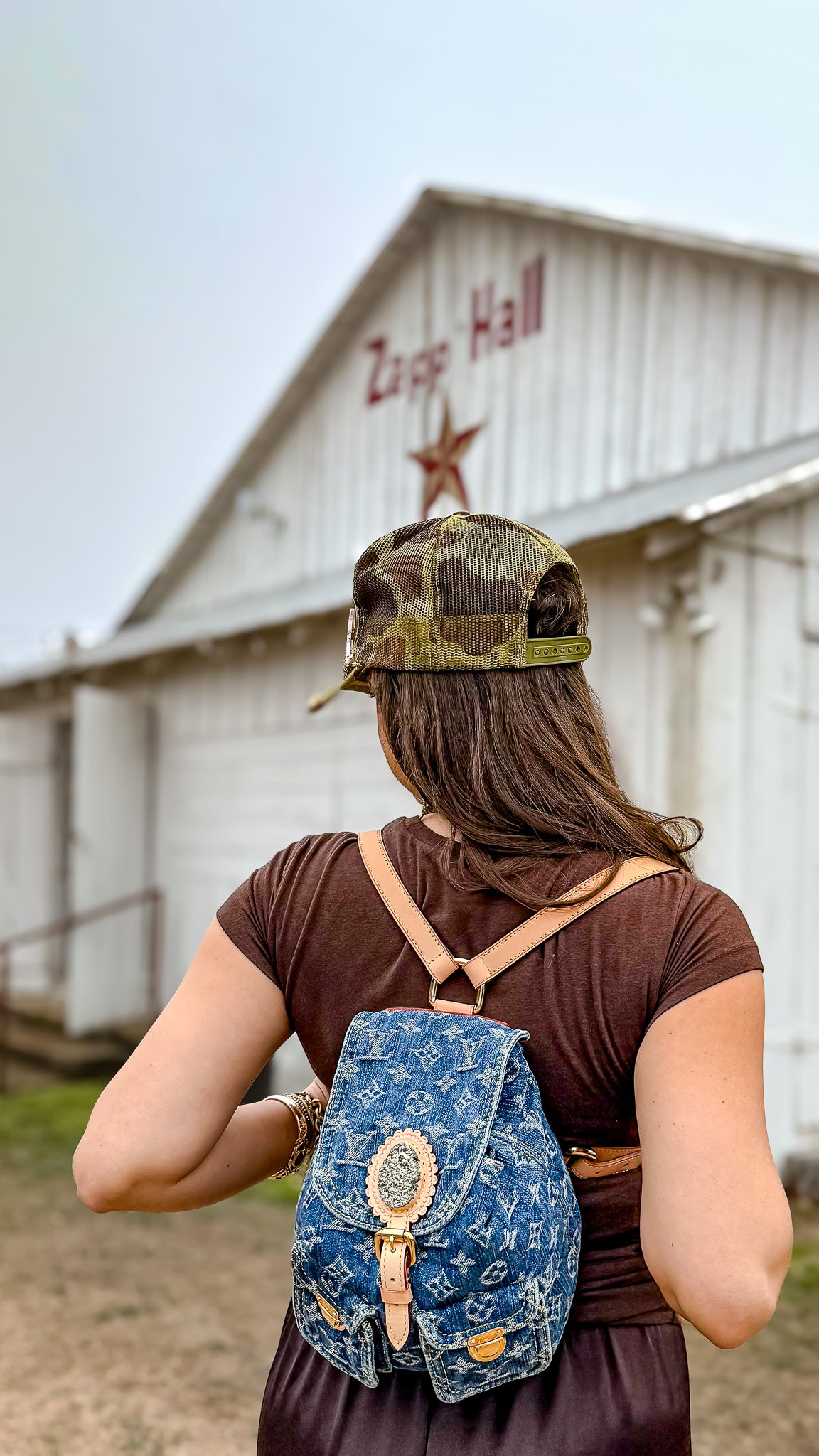 Person wearing a denim backpack and camouflage cap in front of a barn with 'Zapp Hall' sign.