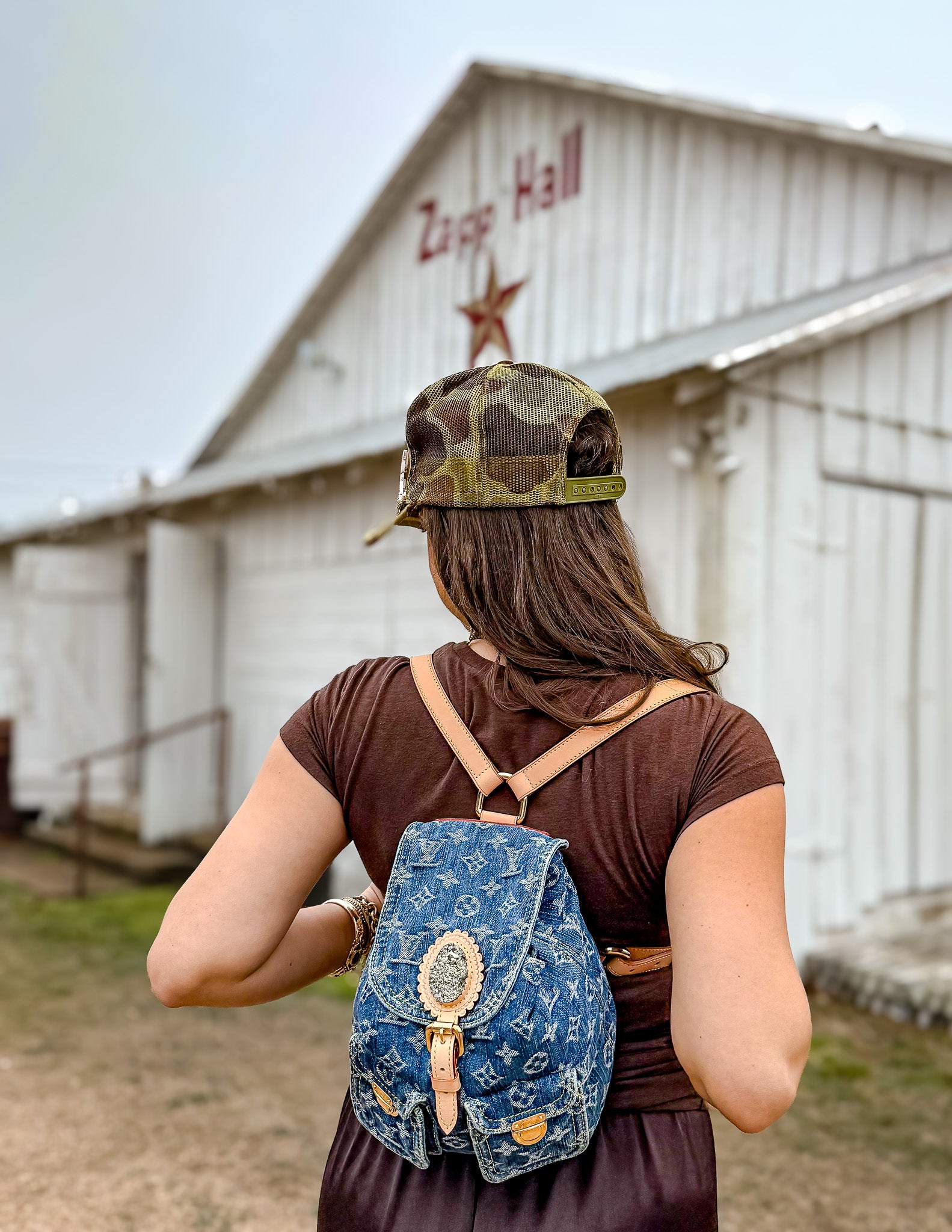 Person wearing a denim backpack and camouflage cap in front of a building with 'Zapp Hall' sign.